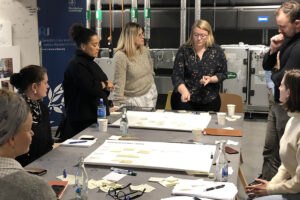Personer som sitter och står runt ett bord och diskuterar. På bordet ligger workshop-material.