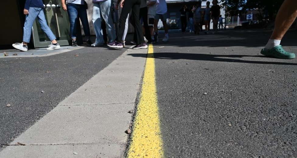 Utomhus, bild på asfalt med en gul linje. I bakgrunden ser man benen på barn som passerar den gula linjen.