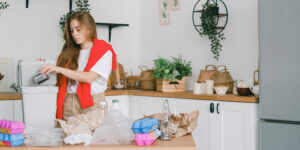Inomhus i ett kök. En kvinna står vid ett bord och sorterar återvinning - metall, plast, äggkartonger och glas.
