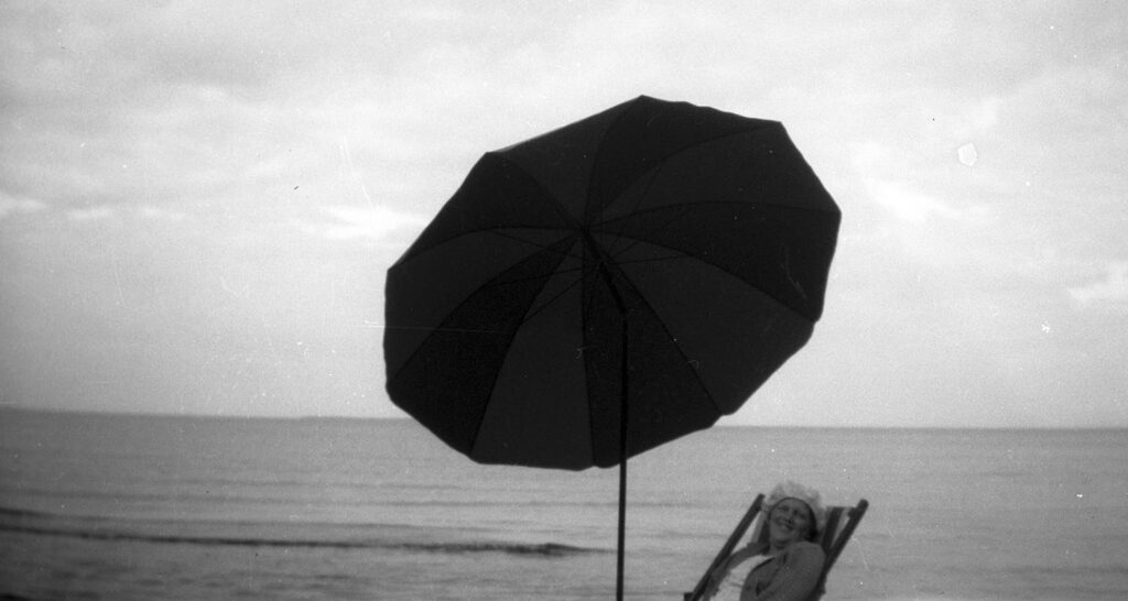 Utomhus. Ett parasoll och överdelen av en solstol där en person sitter. I bakgrunden havet. Svartvitt foto.